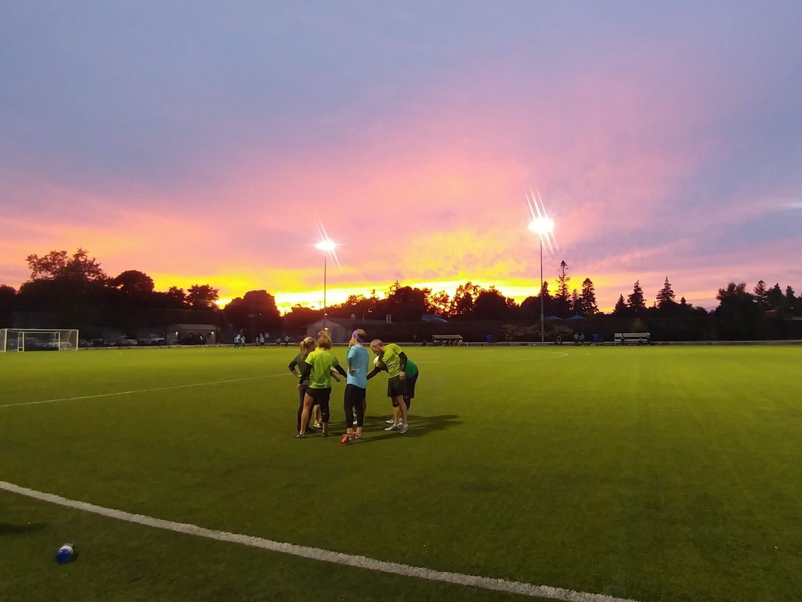 A team circles for a huddle on a turf field at sunset